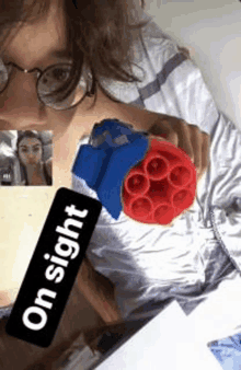 a woman wearing glasses is holding a red and blue toy gun while talking on a video call .