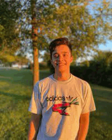 a young man wearing an adidas t-shirt is standing in a park .