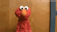 elmo from sesame street is standing in front of a brown wall and looking at the camera .