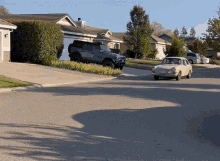 a white car is driving down a street in a residential area with a black suv parked on the side of the road