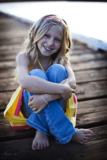 a young girl sits on a wooden dock with her legs crossed and smiles at the camera