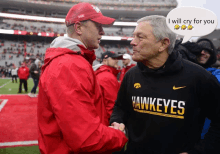 a man wearing a black hawkeyes shirt shakes hands with another man