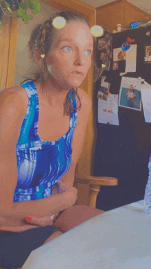 a woman in a blue tank top sits at a table in front of a refrigerator