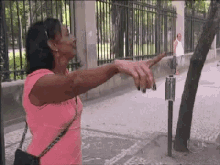 a woman in a pink shirt is standing on a sidewalk with her arms outstretched in front of a fence .