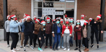 a group of people wearing santa hats and face masks are posing for a picture in front of a building that says happy holidays