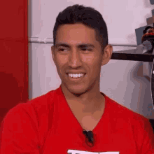 a man in a red shirt is smiling and looking at the camera while sitting in front of a refrigerator .