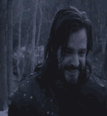 a man with long hair and a beard is standing in the woods .
