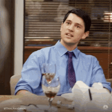 a man in a blue shirt and purple tie is sitting at a table with a glass of wine .