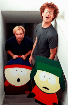 two men standing on a set of stairs holding cardboard cutouts of stan and kyle south park characters