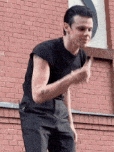 a man in a black shirt and black pants is standing in front of a brick wall giving a thumbs up