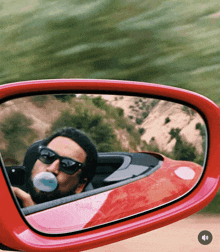 a man blowing a blue bubble in a red car is reflected in the rear view mirror