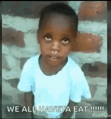 a young boy in a white shirt is standing in front of a brick wall and making a funny face .