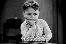 a black and white photo of a little boy sitting at a desk with his hand on his chin and the words `` yesssss '' .