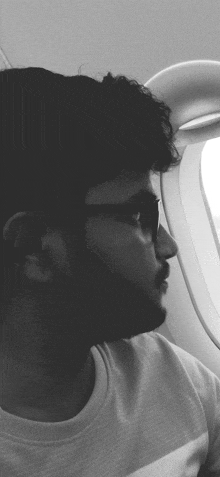 a man wearing glasses and a white shirt looks out a window on an airplane