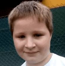 a young boy wearing a white shirt is standing in front of a chain link fence and smiling .