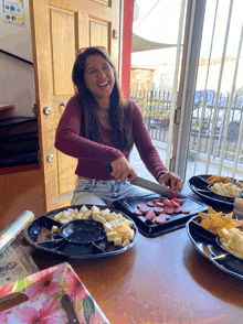 a woman sits at a table with plates of food including a tray of cheese and a tray of sausage