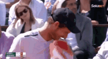 a man in a baseball cap is sitting in a crowd of people watching a tennis match with a scoreboard that says sekabet