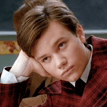 a young man in a red suit and tie is sitting in front of a chalkboard .