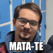 a man wearing glasses and a beard is standing in front of blue lockers with the words mata-te written on his face