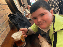 a boy in a green jacket holds a glass of beer with a sticker on it that says " beer "