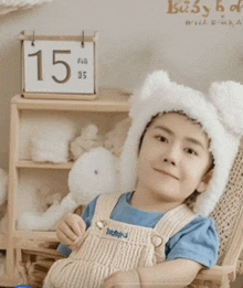 a little boy wearing a white hat and overalls sits in a chair with a calendar that says feb 15 on it