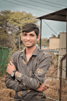 a young man wearing a watch stands with his arms crossed and a barbed wire fence in the background