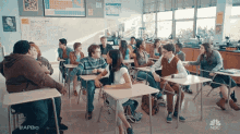 a group of students are sitting at their desks in a classroom with a periodic table on the wall behind them .