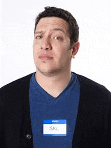 a man wearing a blue shirt and a black cardigan has a name tag on his neck .