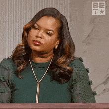 a woman wearing a green top and a gold necklace is sitting in front of a sign that says be