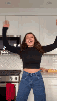 a woman in a black crop top and blue jeans is dancing in a kitchen with her arms outstretched .