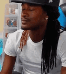 a man with dreadlocks wearing a hat and a white shirt is sitting down with his eyes closed .