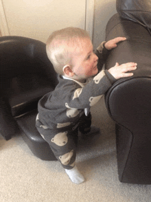 a baby wearing bear pajamas reaches for a couch arm