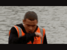 a man wearing an orange vest and a black shirt is standing in front of a body of water .