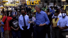 a man in a blue shirt and tie stands in front of a crowd with a blm sign behind him