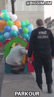 a man in a black uniform with the word straz miejska on the back walks past a bunch of balloons