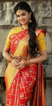 a woman wearing a red and yellow saree is standing in front of a stone wall .