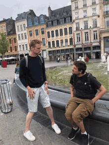 two men standing and sitting in front of a fountain in front of a building that has a sign that says ' boutique '