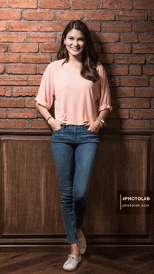 a woman in a pink shirt and blue jeans is standing in front of a brick wall with her hands in her pockets