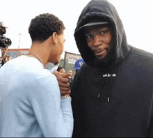 a man in a hoodie talks to another man in a light blue shirt