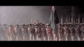 a group of soldiers marching in a line with guns and a flag in the background .