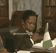 a young boy in a suit and bow tie is sitting at a desk with a flag on it .