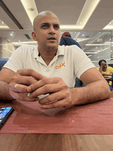 a man wearing a white shirt with gsk on the front sits at a table