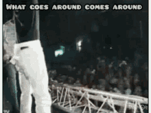 a man in white pants is standing on a stage in front of a crowd with the words `` what goes around comes around '' .