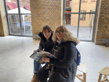 two women are sitting in front of a brick wall and one is reading a magazine