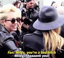 a woman in a hat is talking to another woman in a crowd while wearing sunglasses and a leather jacket .