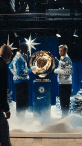 a man in a blue sweater holds a trophy with the chelsea logo on it