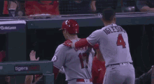 two baseball players are standing next to each other in a dugout . one of the players has the number 4 on his jersey .