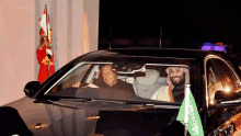 two men are sitting in a car with a saudi flag hanging from the windshield