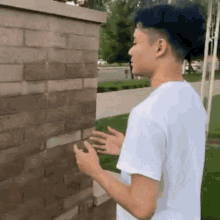 a young man in a white shirt is standing in front of a brick wall and looking at his phone .