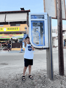 a boy in a nasa shirt stands in front of a phone booth on a street